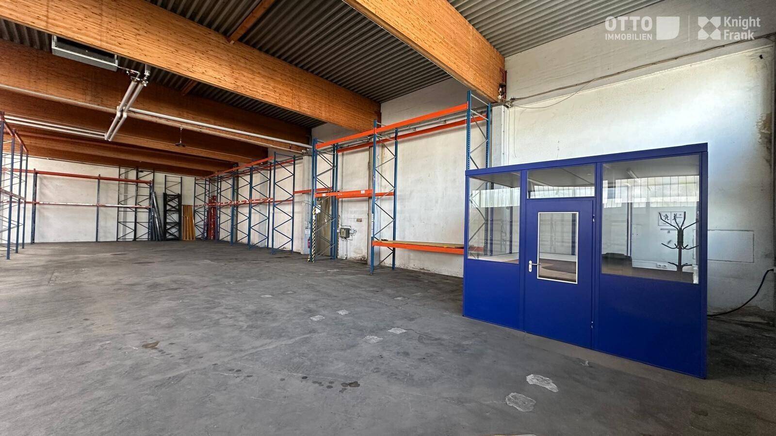 Industrielle Lagerhalle mit Betonboden und einer blauen Bürobox, umgeben von Regalsystemen.