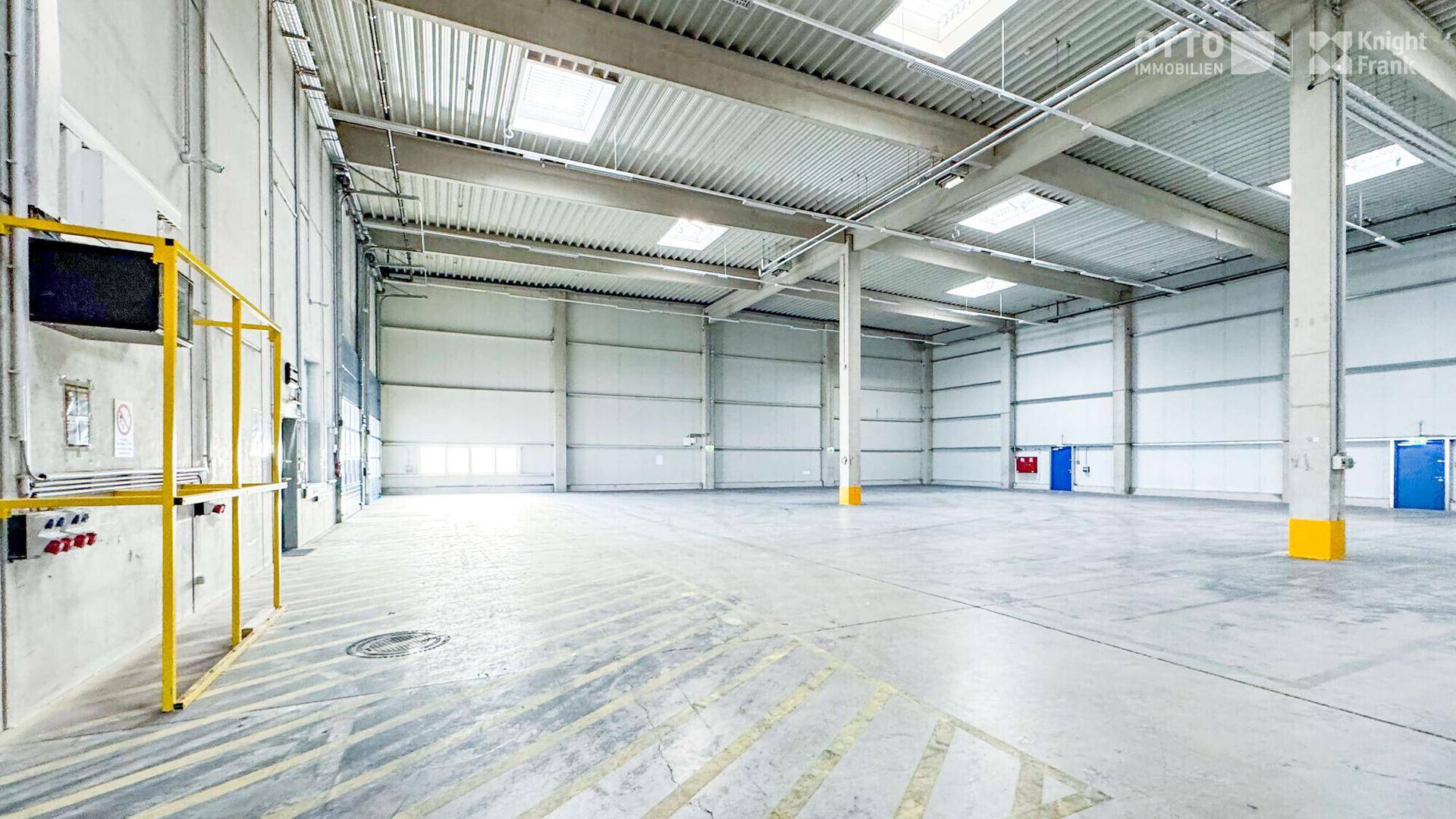 Weitläufige, leere Lagerhalle mit hohen Decken, Oberlichtern und einem robusten Betonboden.