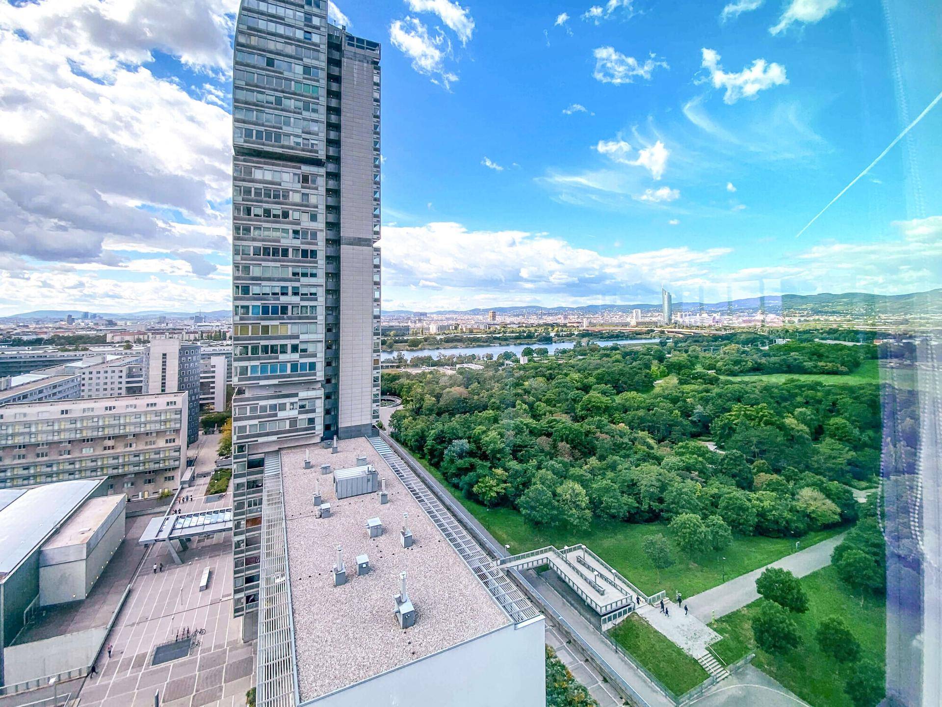 Panoramablick auf ein modernes Hochhaus, umgeben von Grünflächen und einem Fluss, unter blauem Himmel.