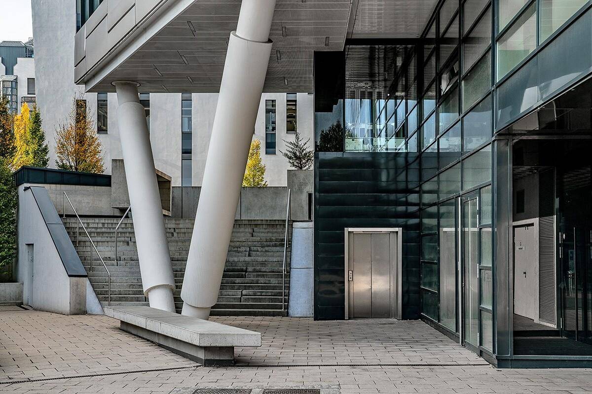 Moderner Gebäudeeingang mit Aufzug, Treppen und großen Glasflächen, umgeben von Betonstrukturen.