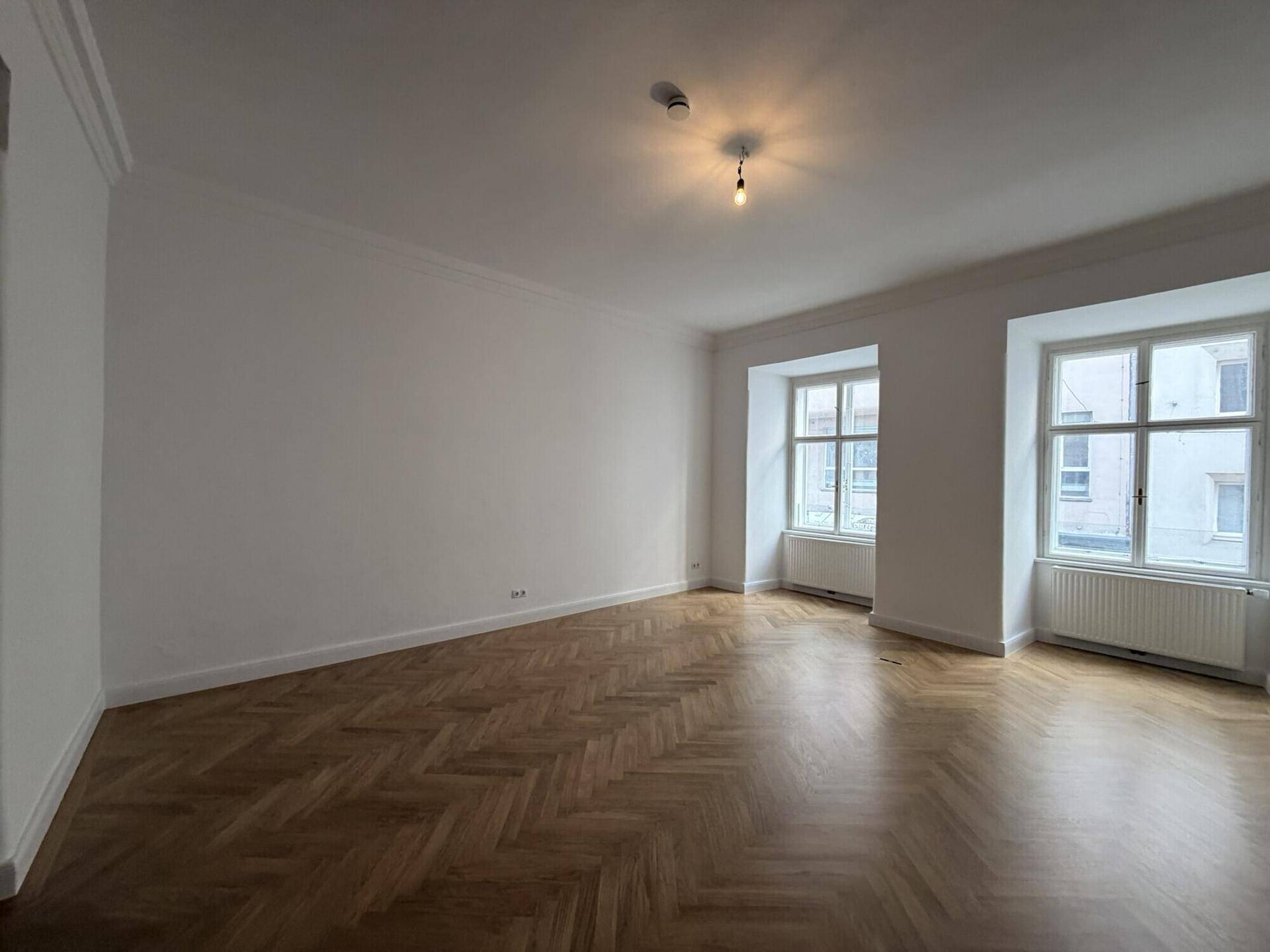 Helles Wohnzimmer mit traditionellem Fischgrätparkett und hohen Wänden, die eine geräumige Atmosphäre schaffen.