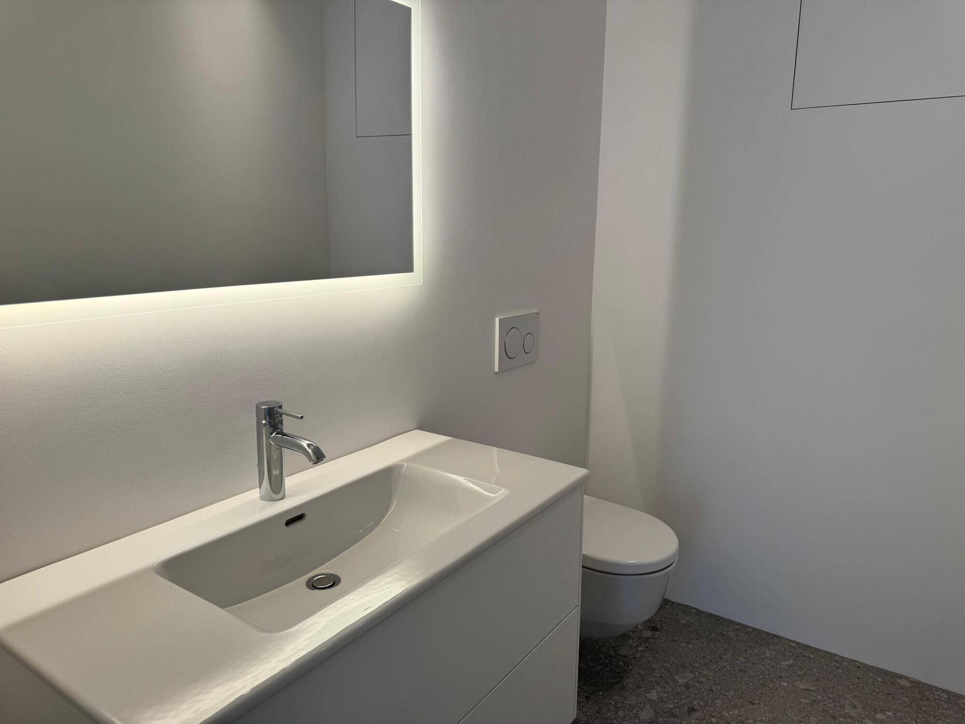 Modernes Badezimmer mit weißem Waschtisch, integriertem Spiegel mit Beleuchtung und wandhängendem WC in minimalistischem Design.