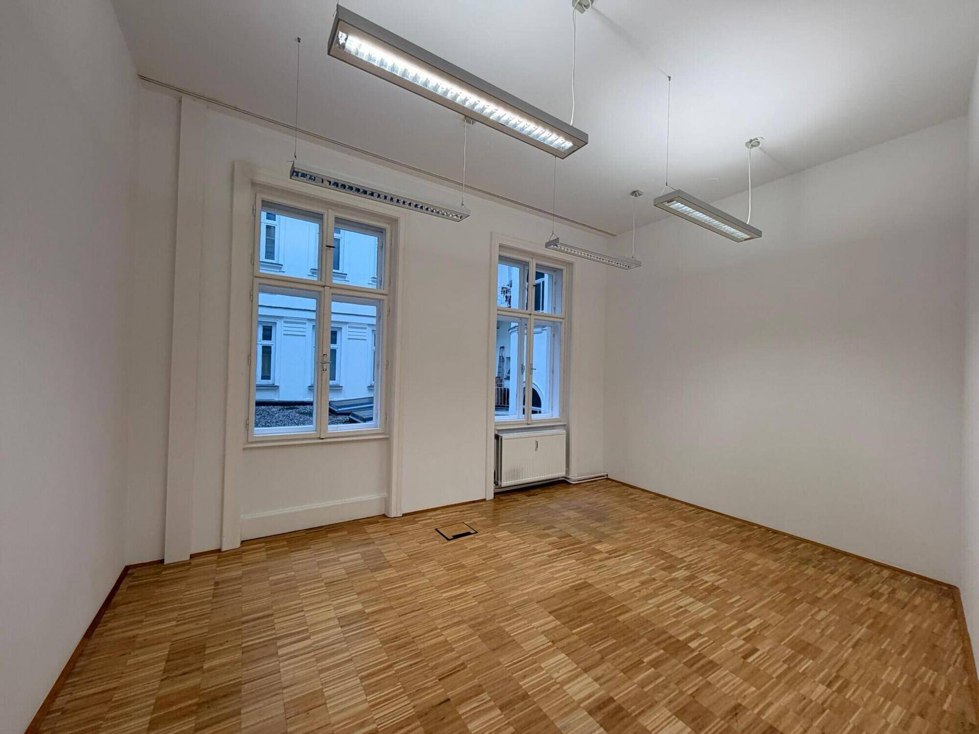 Heller Büroraum mit Parkettboden, zwei großen Fenstern und moderner Deckenbeleuchtung, bereit für individuelle Gestaltung.