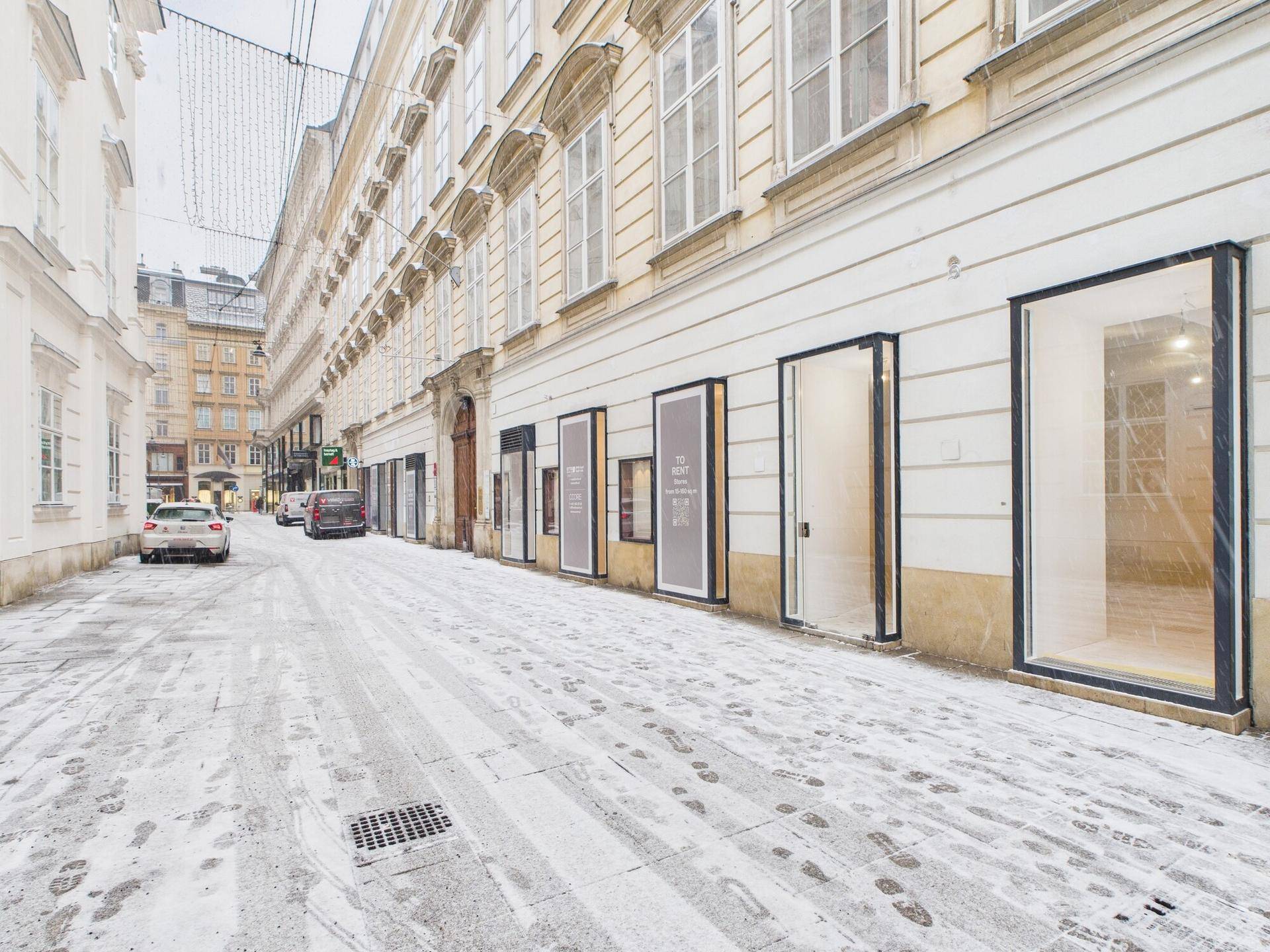 Schneebedeckte Straße vor einem historischen Gebäude mit modernen Ladenfronten im Stadtzentrum.