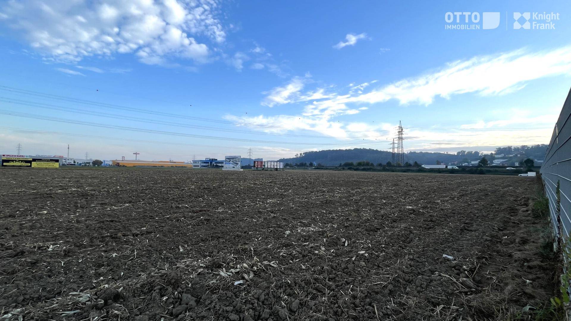 Gepflügtes Feld mit Strommasten und Industriegebäuden in der Ferne, begrenzt durch einen Zaun.