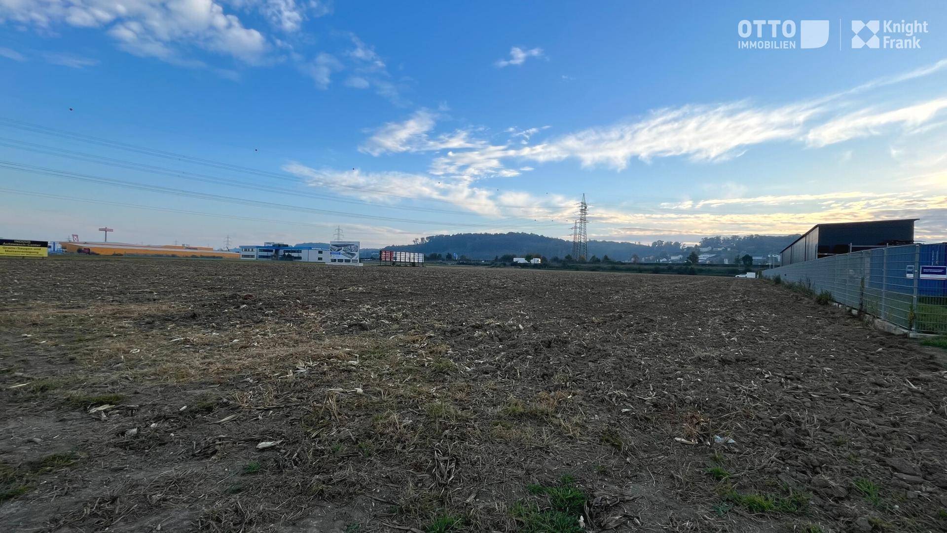 Weites, gepflügtes Feld mit Strommasten und Gewerbegebäuden am Horizont, begrenzt durch einen Zaun.