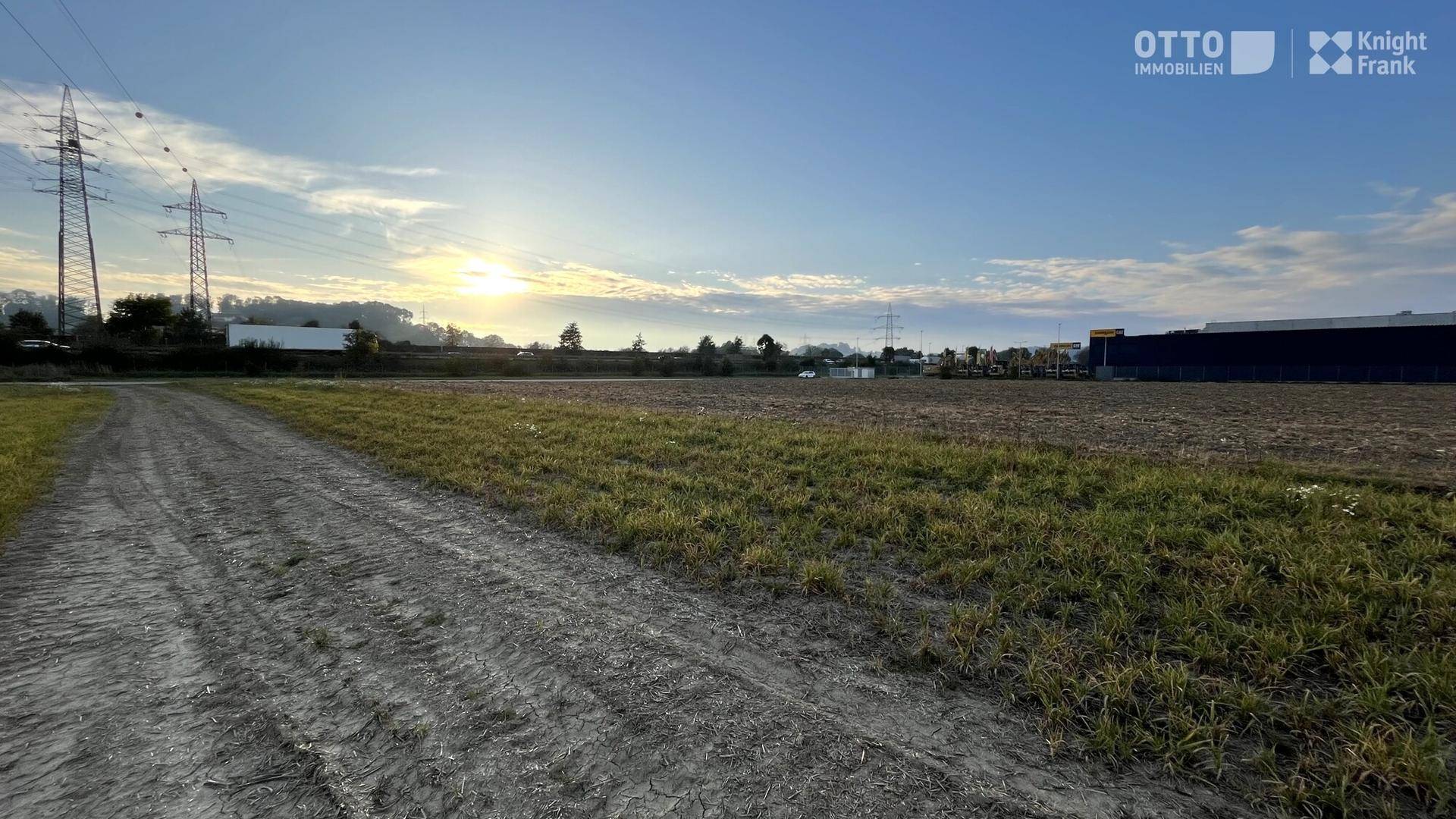 Sonnenaufgang über einem weiten Feld mit Strommasten und entfernten Gebäuden, ein Feldweg führt hindurch.