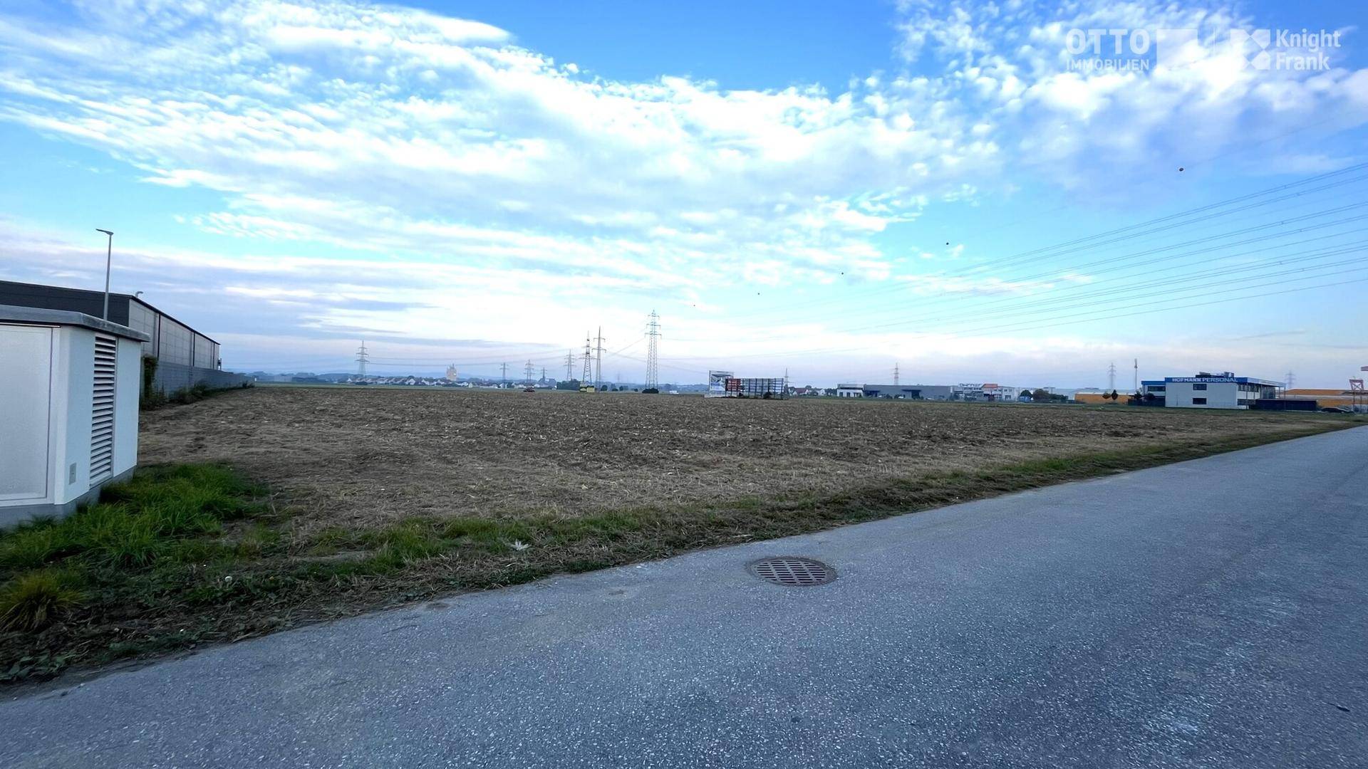 Blick über ein weites Feld neben einer Straße mit Kanaldeckel, Strommasten und Gebäuden in der Ferne.