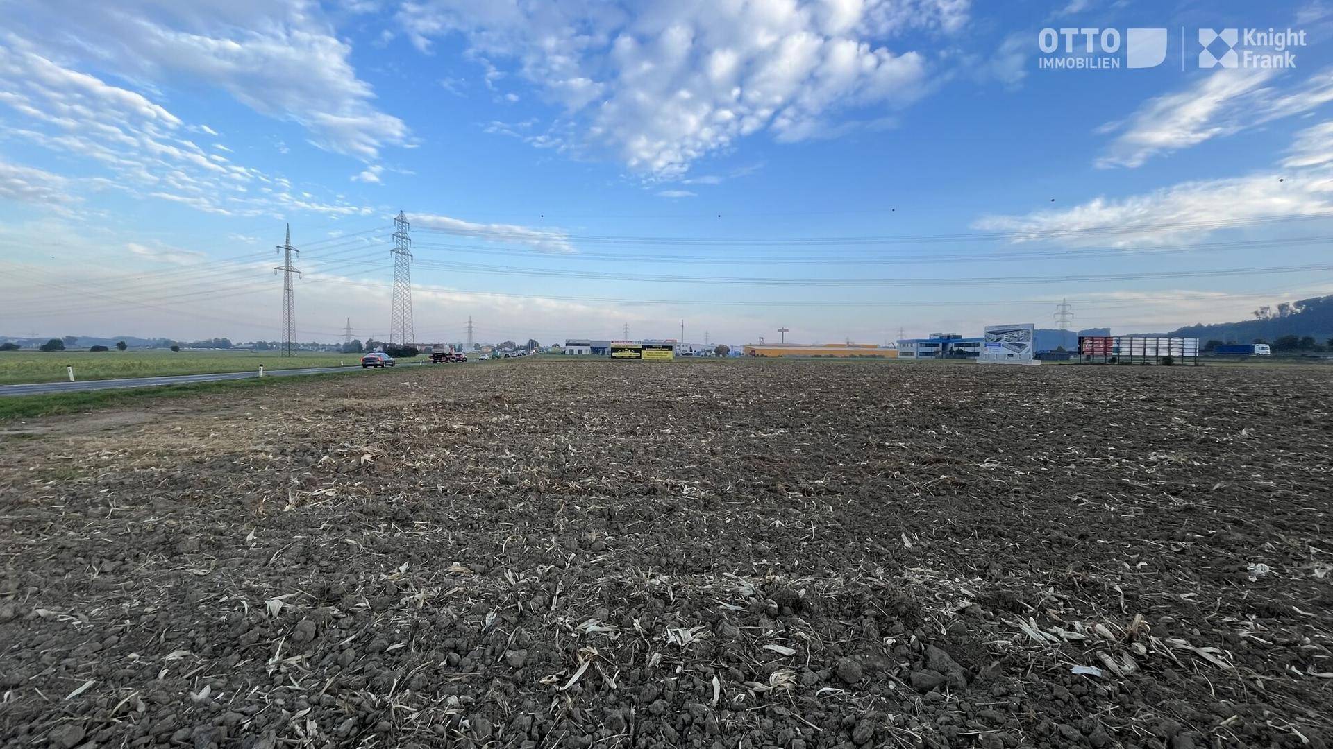 Gepflügtes Feld mit Strommasten und Industriegebäuden in der Ferne unter einem bewölkten blauen Himmel.