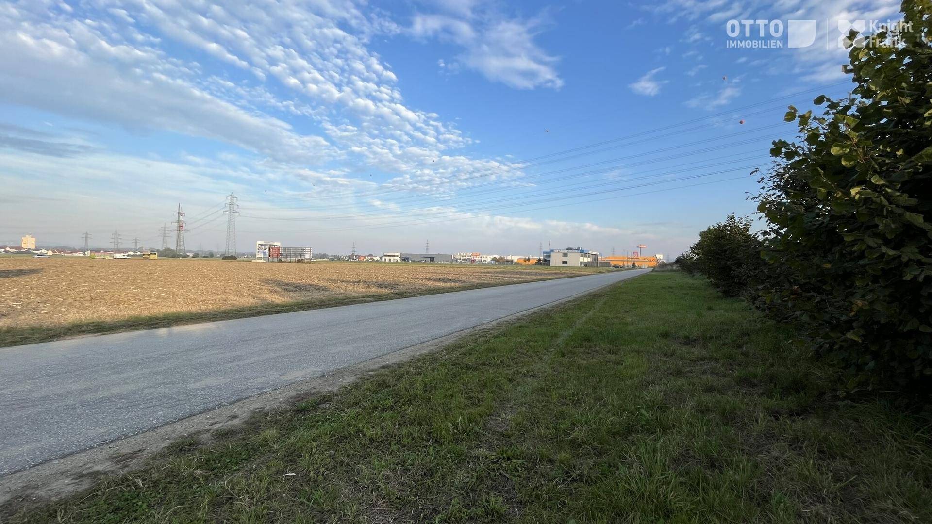 Blick über ein weites Feld mit einer Straße, Strommasten und Gebäuden in der Ferne unter klarem Himmel.