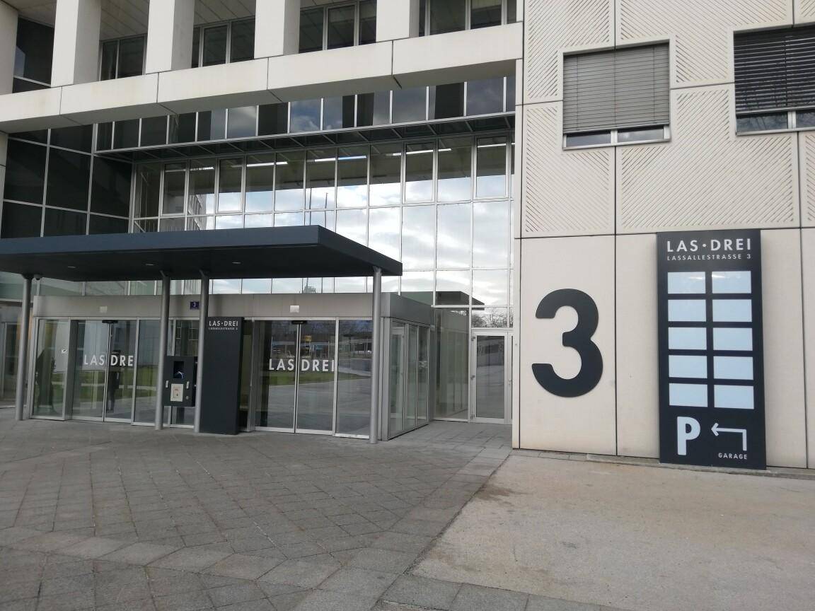 Gebäudeeingang mit großen Glasfronten, einem Vordach und Hinweisschildern zur Tiefgarage.