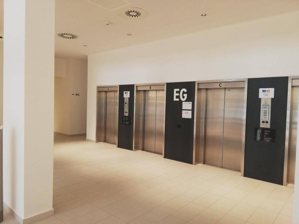 Heller Flur im Erdgeschoss mit mehreren modernen Aufzügen aus Edelstahl und Fliesenboden.