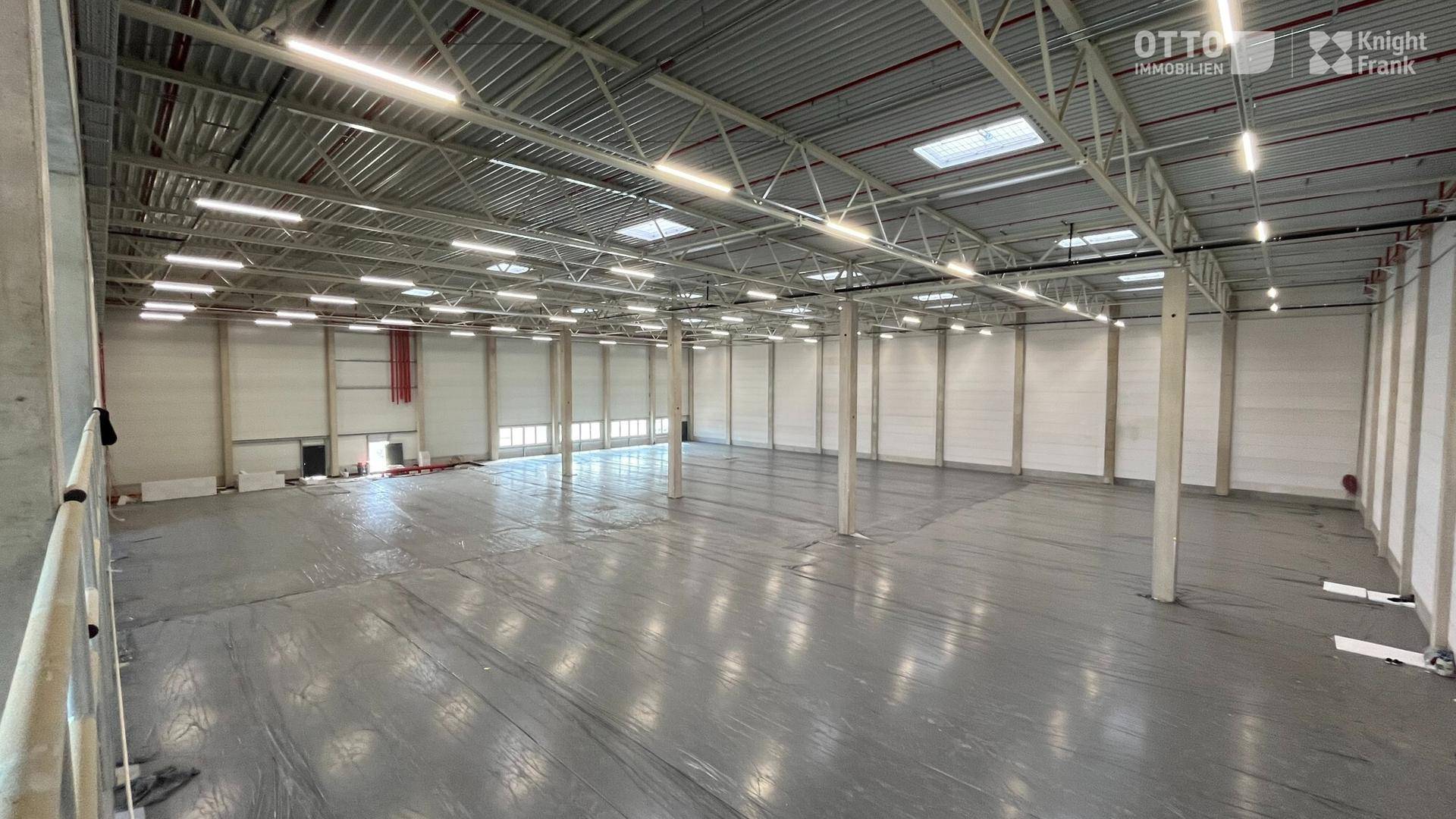 Geräumige, leere Lagerhalle mit hoher Decke, vielen Leuchten und Oberlichtern für Tageslicht.