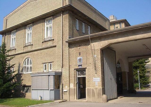 Historisches Backsteingebäude mit markanten Fenstern und einem überdachten Eingangsbereich, umgeben von Grün.