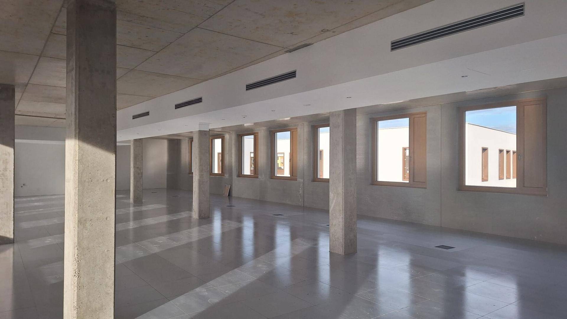 Weitläufiger, unmöblierter Bürobereich mit Betonpfeilern, vielen Fenstern und Lüftungsschlitzen an der Decke.
