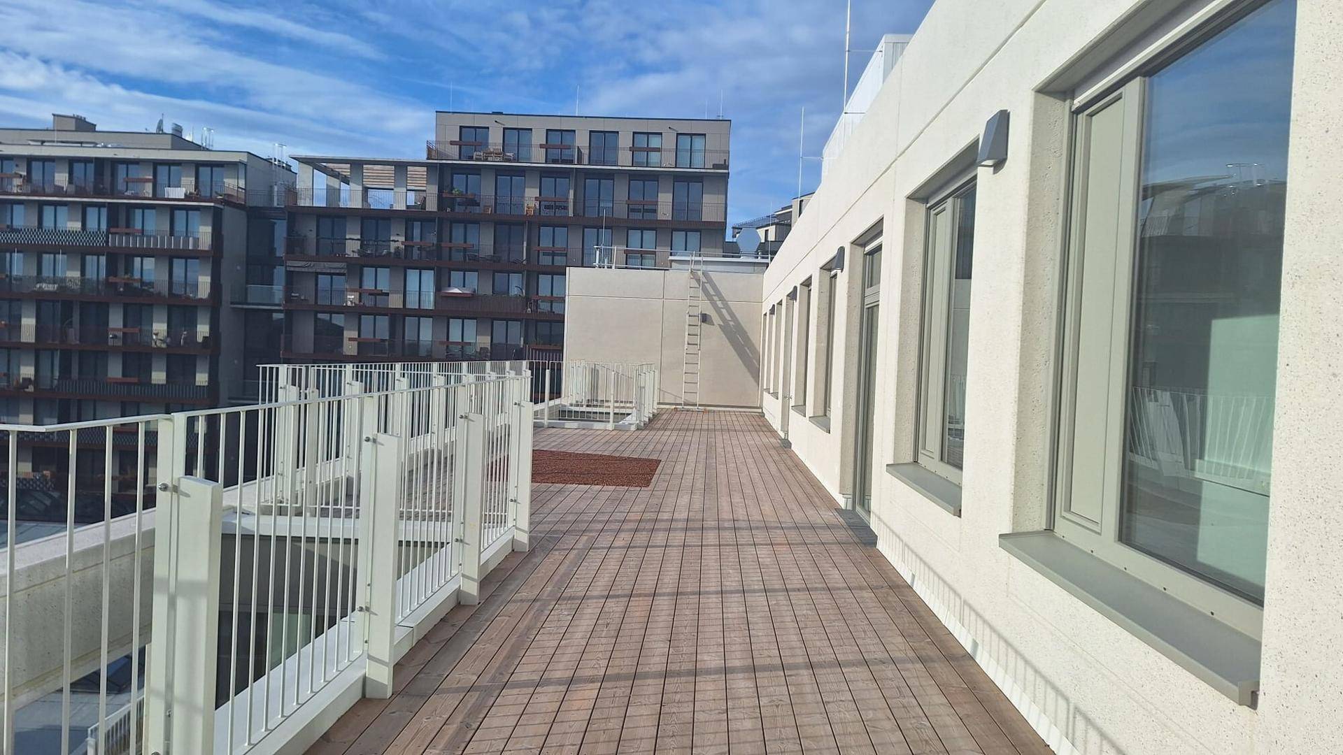 Weitläufige Dachterrasse mit Holzdielenboden und weißem Geländer, angrenzend an die moderne Gebäudefassade.
