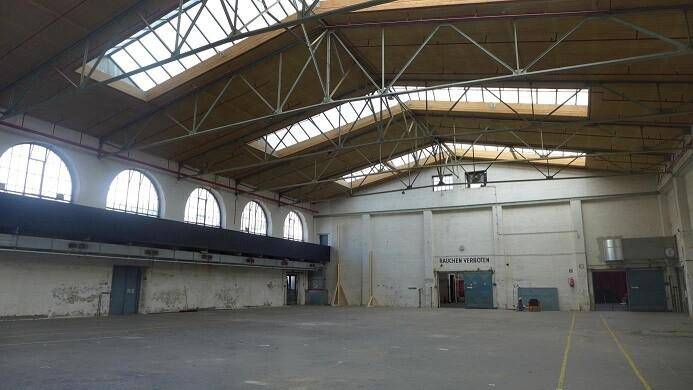 Weitläufige Industriehalle mit gewölbter Dachkonstruktion, Oberlichtern und großen Bogenfenstern.