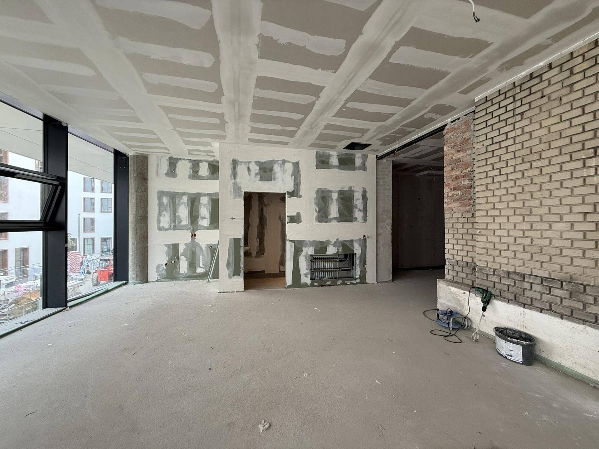 Innenraum im Rohbau mit unverputzten Ziegelwänden, Trockenbau und großen Fenstern.