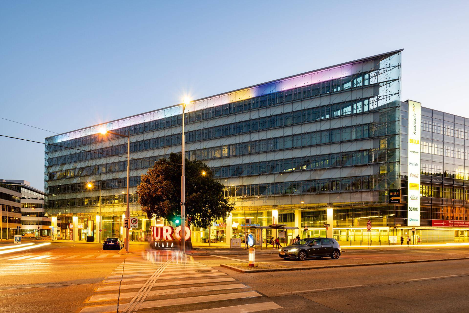 Weitläufige Ansicht des modernen Bürokomplexes "EURO PLAZA" mit beleuchteter Glasfassade und städtischer Umgebung bei Dämmerung.