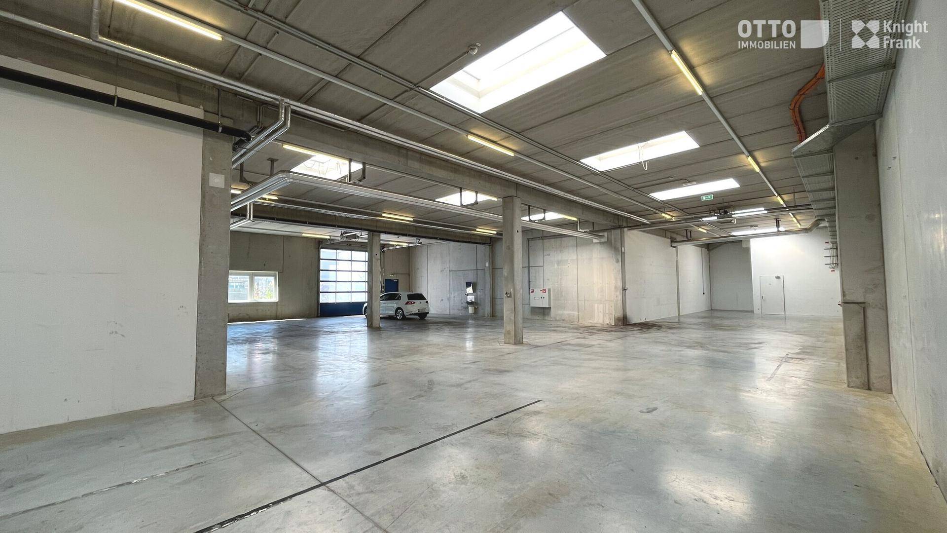 Große Lagerhalle mit Betonboden, hohen Decken und einem Rolltor im Hintergrund, ein Auto ist sichtbar.
