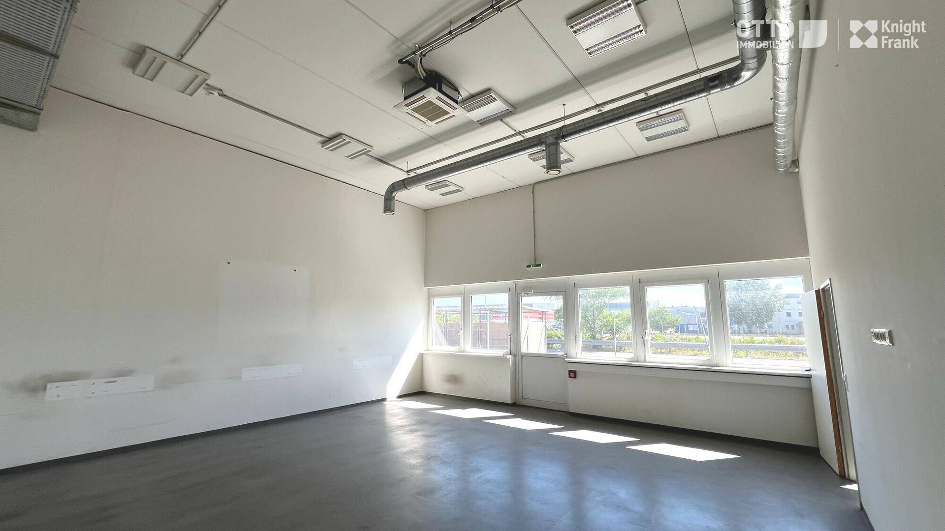 Heller Raum mit hohen Decken, dunklem Boden und einer Fensterfront mit Zugang nach draußen.