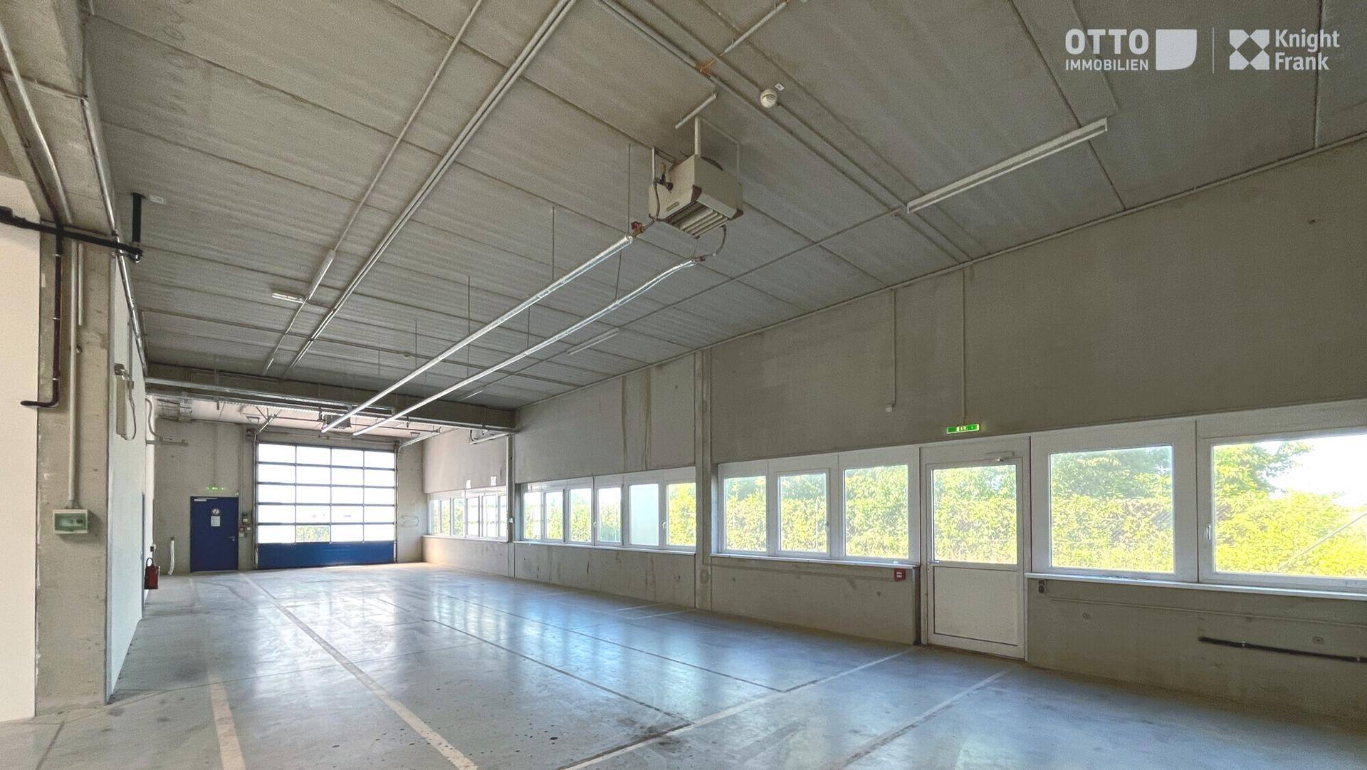 Helle Lagerhalle mit Betonboden, einer Reihe von Fenstern und einem großen Rolltor am Ende des Raumes.