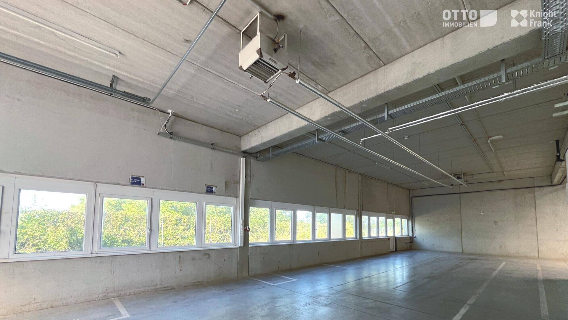 Großer, leerer Lagerraum mit Betonboden, hohen Decken und einer Reihe von Fenstern für Tageslicht.