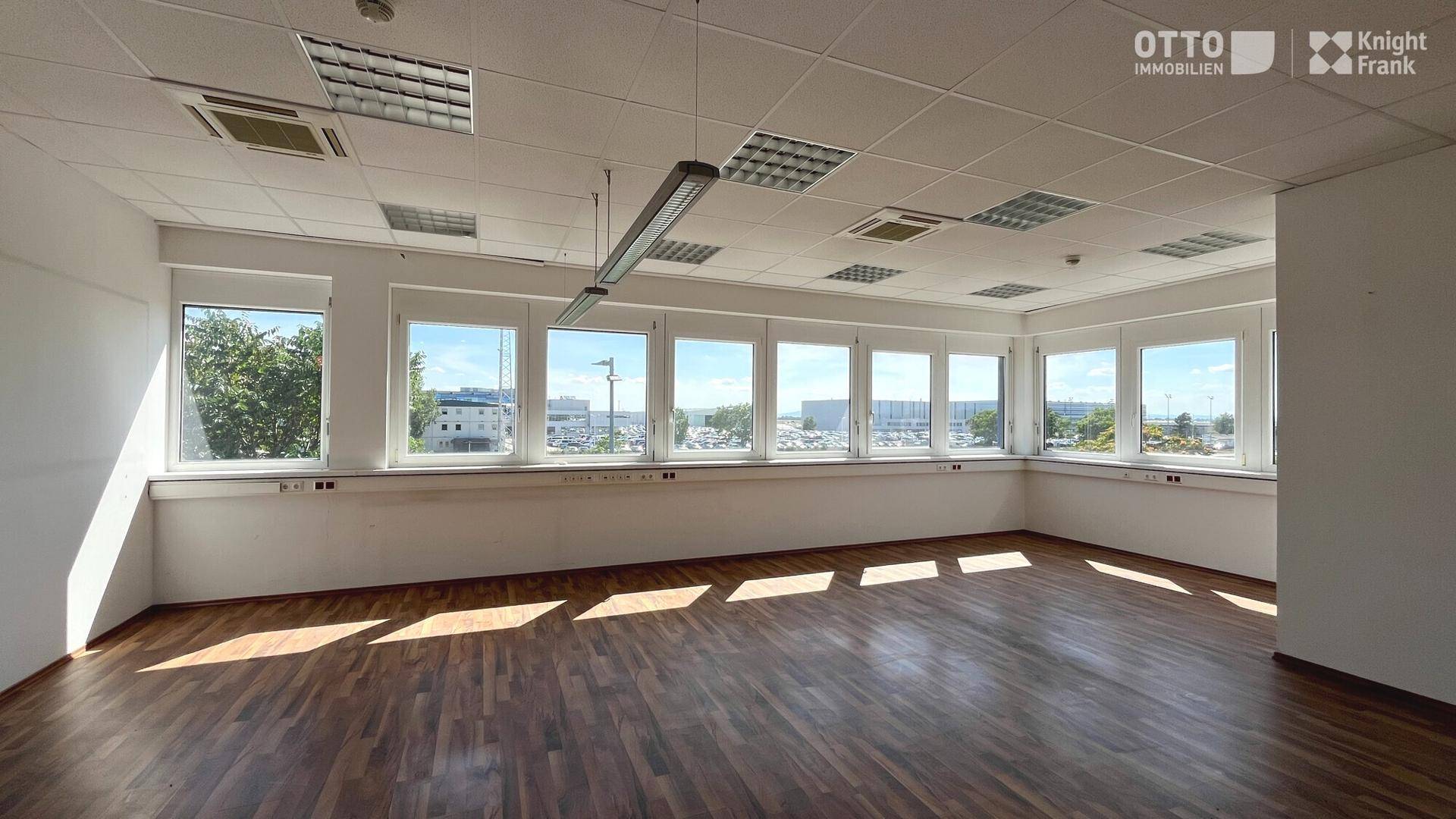 Helles Büro mit Holzfußboden, abgehängter Decke und großen Fenstern mit Blick auf die Umgebung.