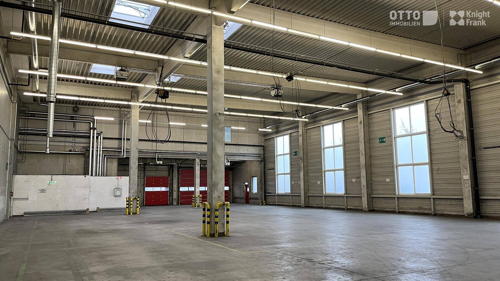 Weitläufige, leere Lagerhalle mit hohen Decken, großen Fenstern und sichtbaren Versorgungseinrichtungen.