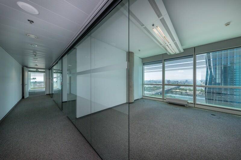 Heller Büroflur mit Glaswänden, der zu weiteren Büroräumen mit Stadtblick führt.