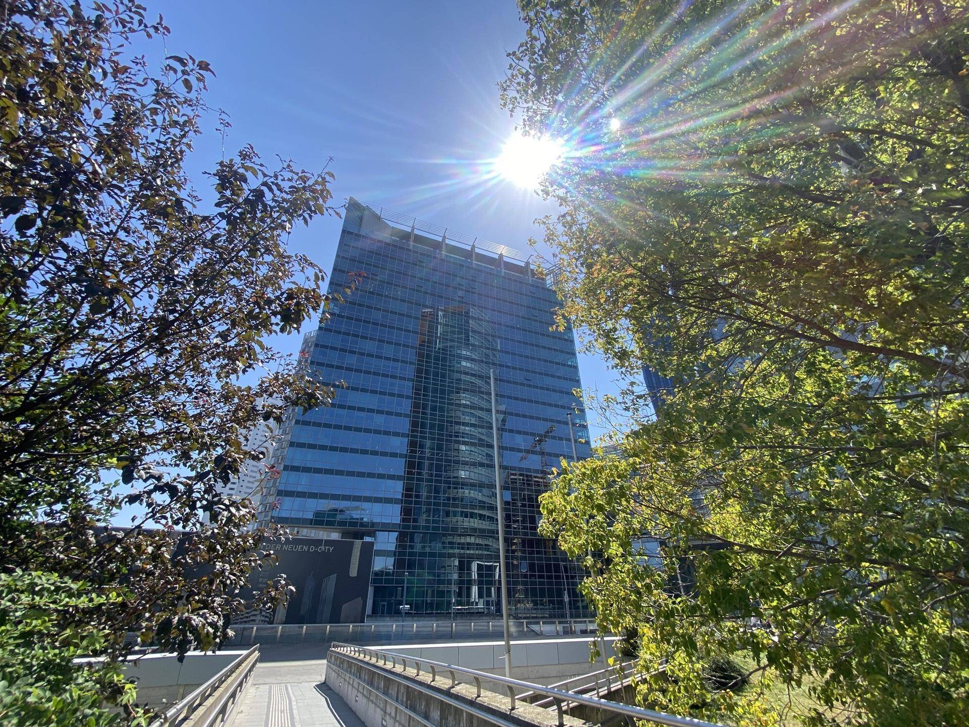 Modernes Hochhaus mit Glasfassade, umgeben von Bäumen, unter strahlendem Sonnenschein.