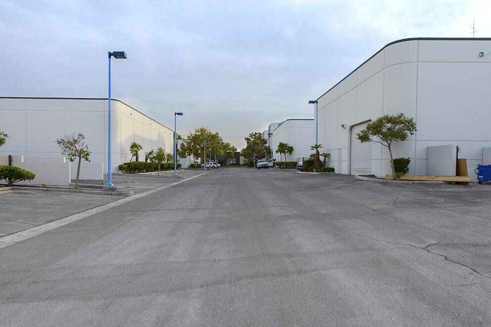Zufahrtsstraße zwischen mehreren weißen Gewerbegebäuden mit Bäumen und Laternen in einem Industriepark.