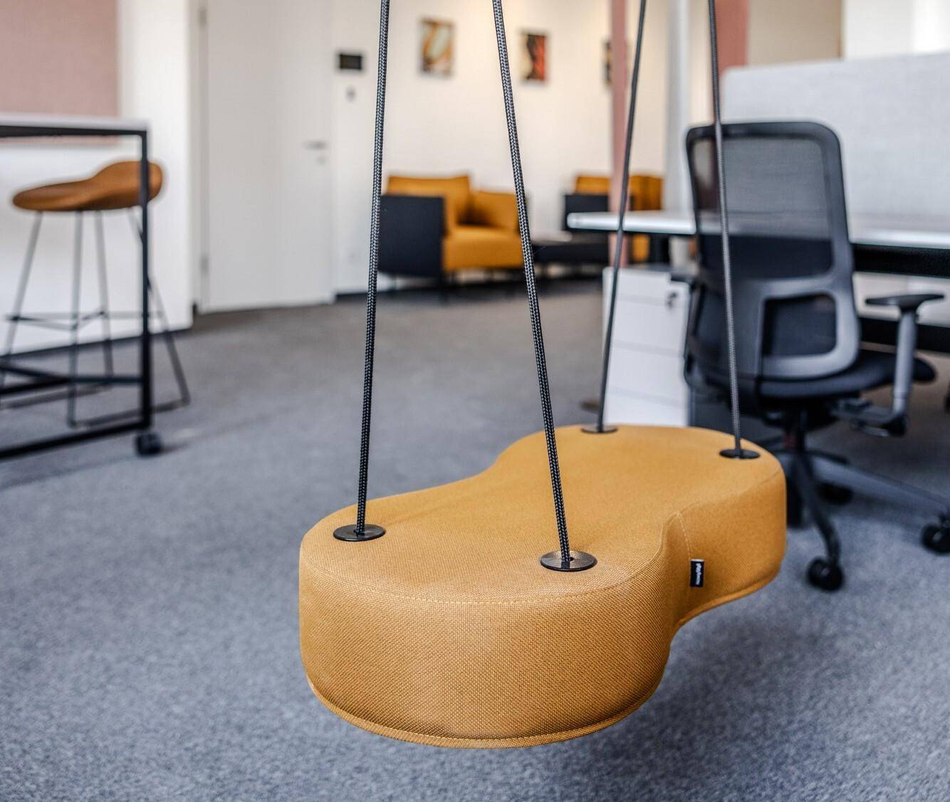 Detailaufnahme einer modernen Schaukel im Bürobereich mit Teppichboden und weiteren Möbeln im Hintergrund.