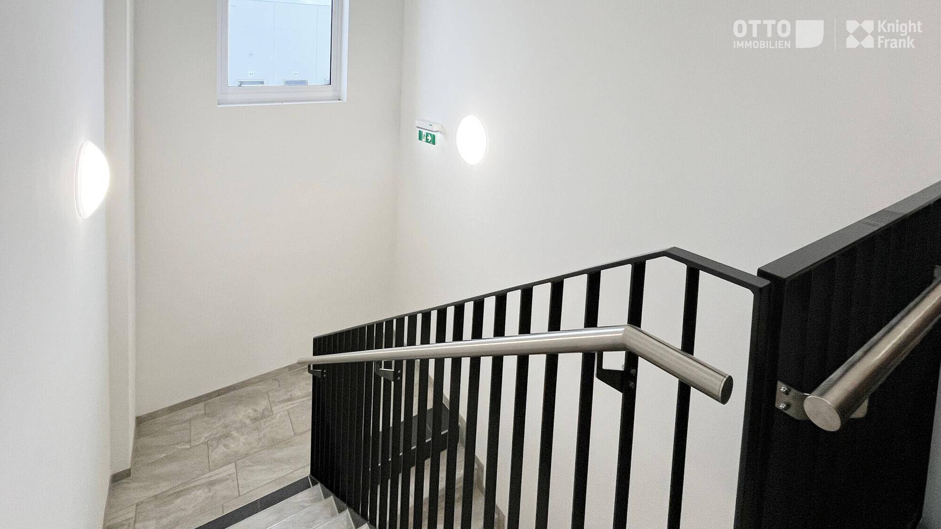 Modernes Treppenhaus mit Metallgeländer, Fliesenboden und einem Fenster für natürliches Tageslicht.