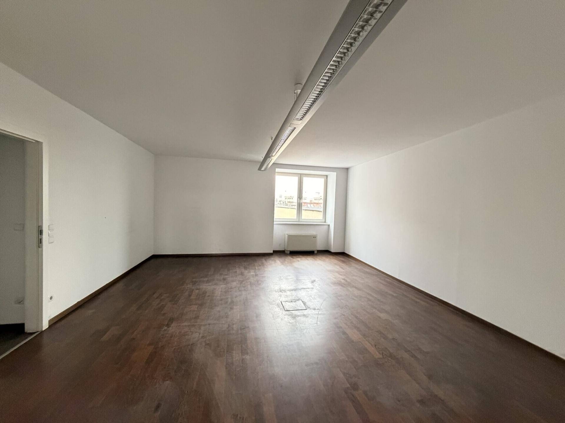 Langer, leerer Raum mit dunklem Holzboden, weißen Wänden und einem Fenster, das Tageslicht hereinlässt.