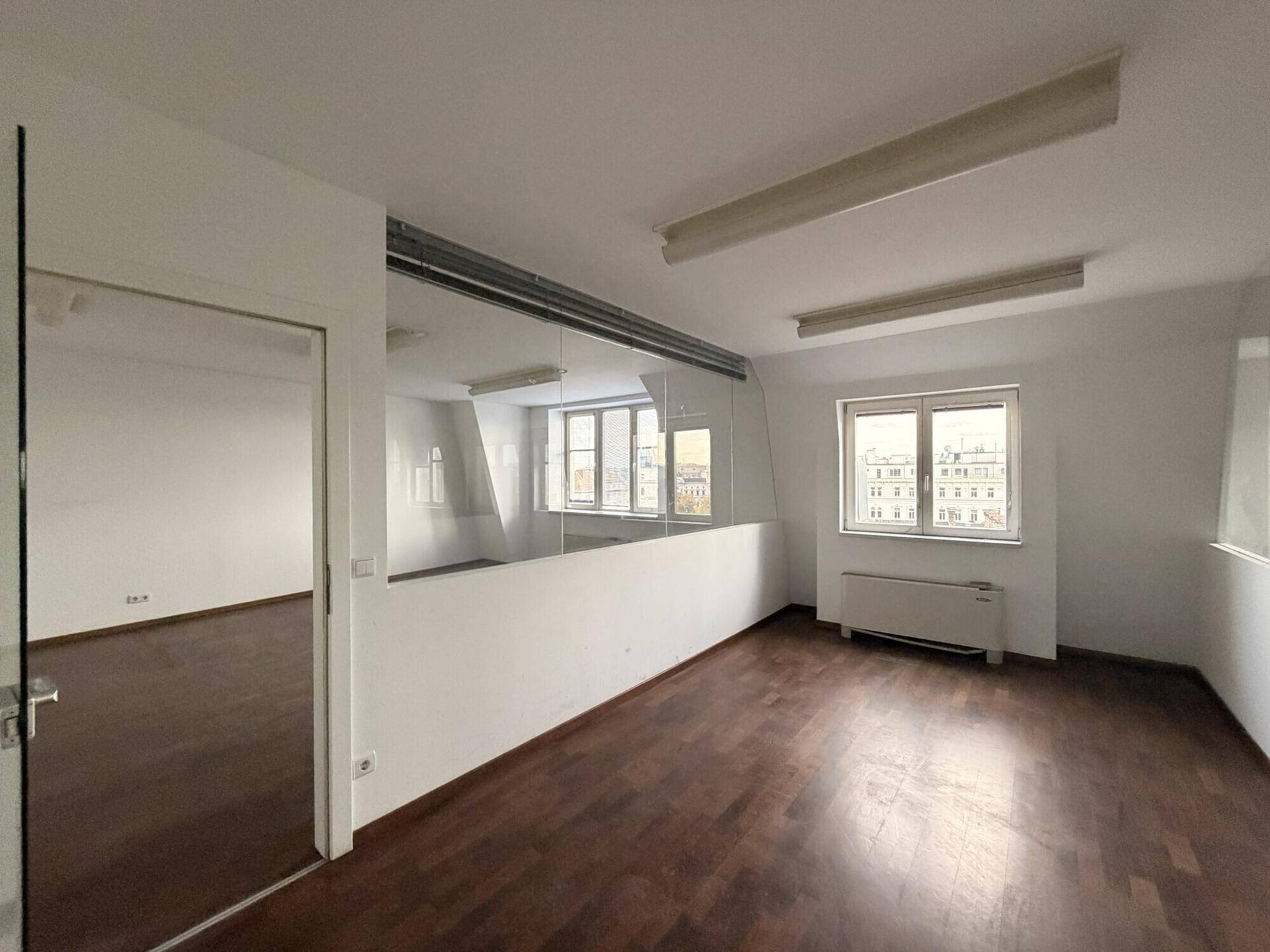 Ansicht des Büroraums mit dunklem Holzboden, weiß gestrichenen Wänden und einer Glastür sowie Glasabtrennung.
