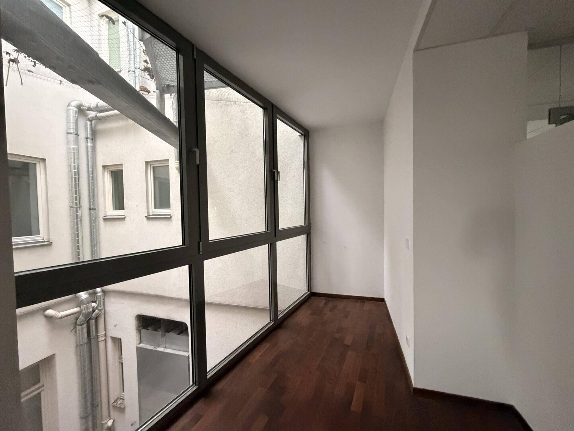 Raum mit großer Fensterfront zum Innenhof, dunklem Holzboden und weißen Wänden, gut beleuchtet.