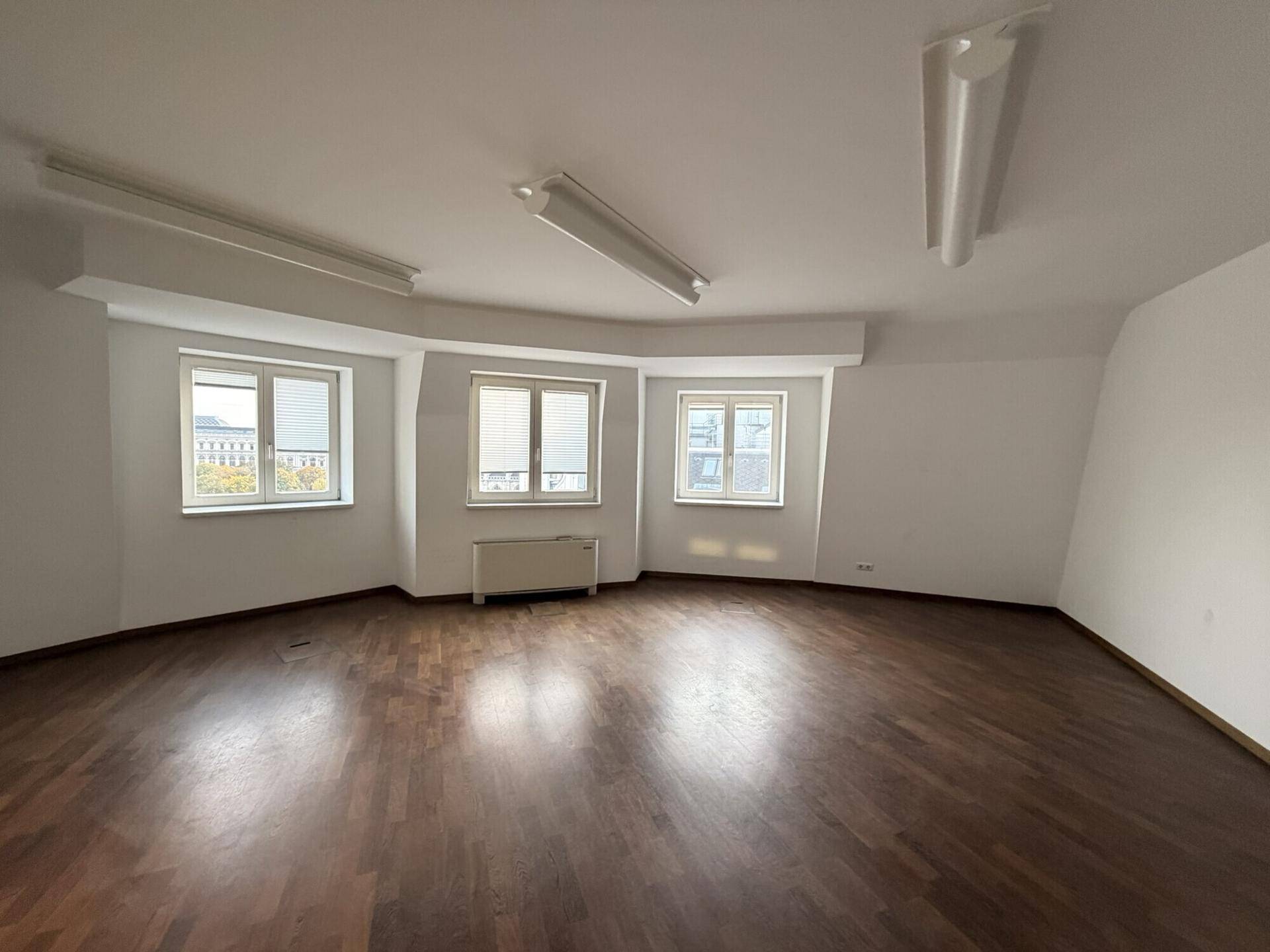 Heller, leerer Raum mit dunklem Holzboden, weißen Wänden und drei Fenstern für viel Tageslicht.