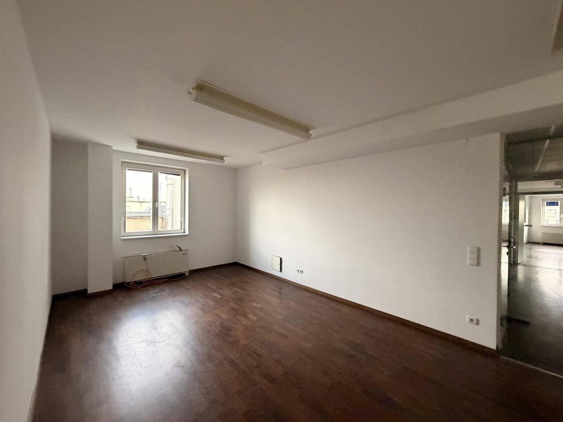 Heller, leerer Raum mit dunklem Holzboden, weißen Wänden und einem Fenster, das Tageslicht spendet.