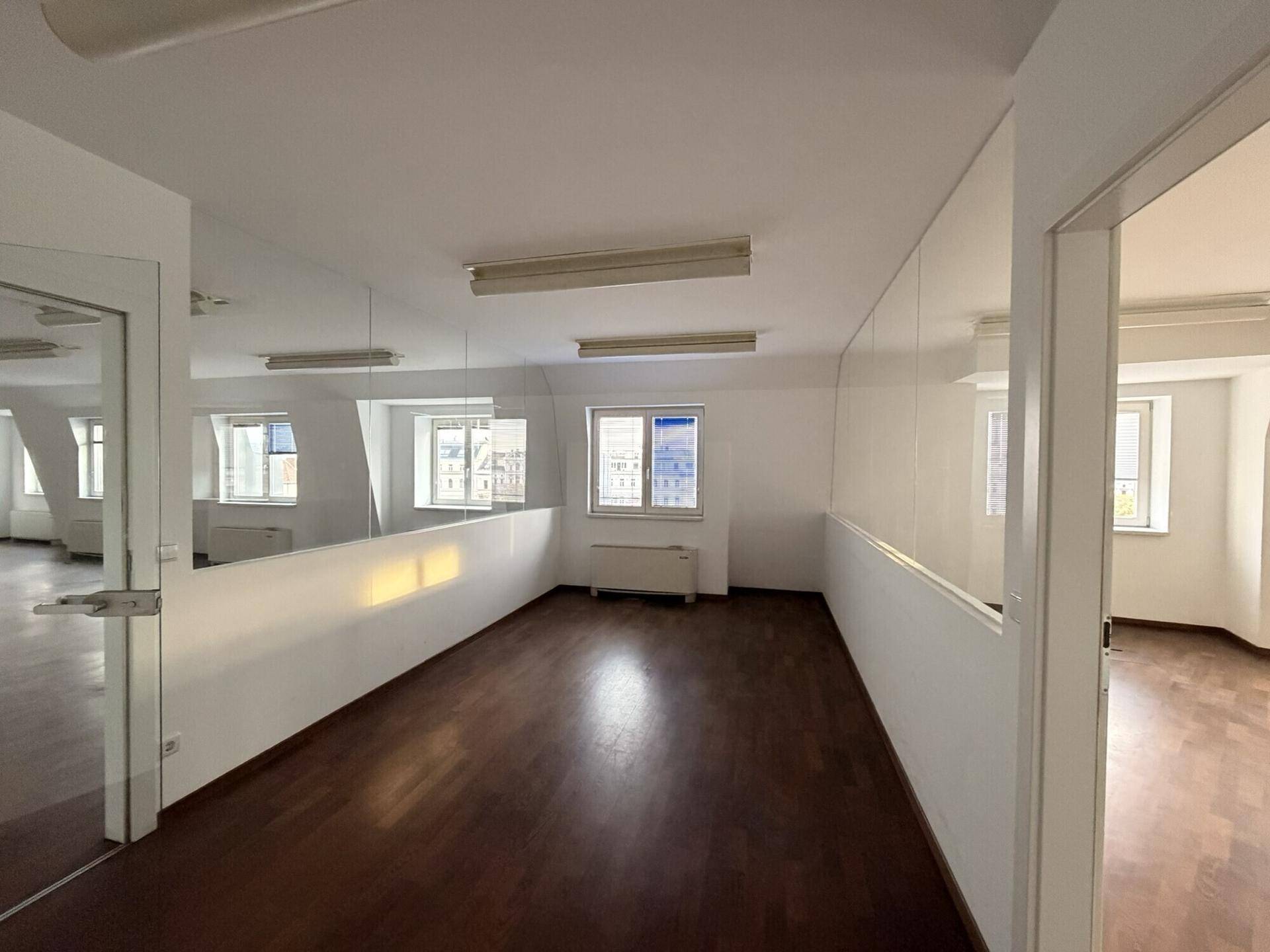 Geräumiger Bürobereich mit dunklem Holzboden, weißen Wänden und einer modernen Glasabtrennung.