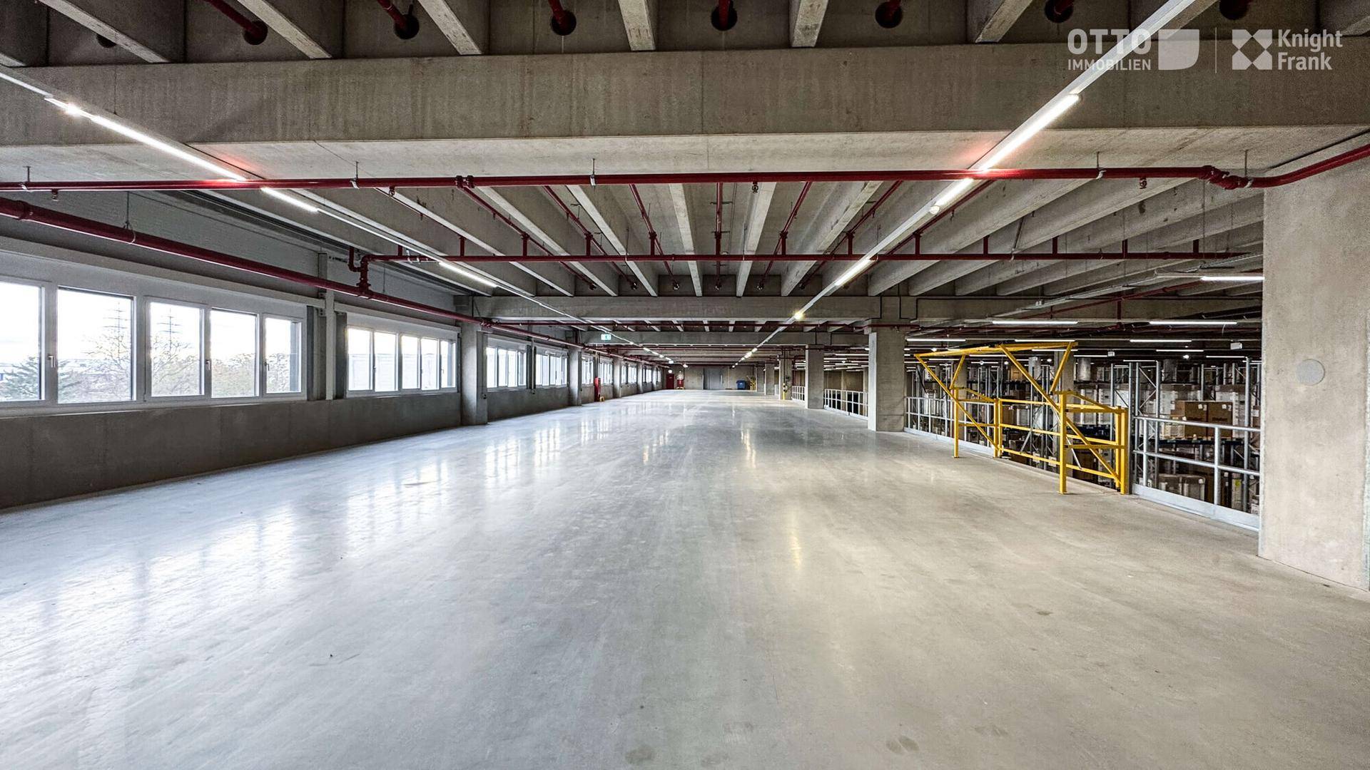 Große, leere Lagerfläche mit vielen Fenstern, die viel Tageslicht hereinlassen und roten Rohren.