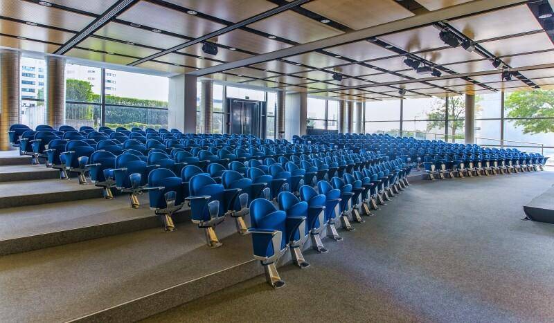Großer Hörsaal mit vielen blauen Sitzen, einer Bühne und großen Fenstern, ideal für Präsentationen.