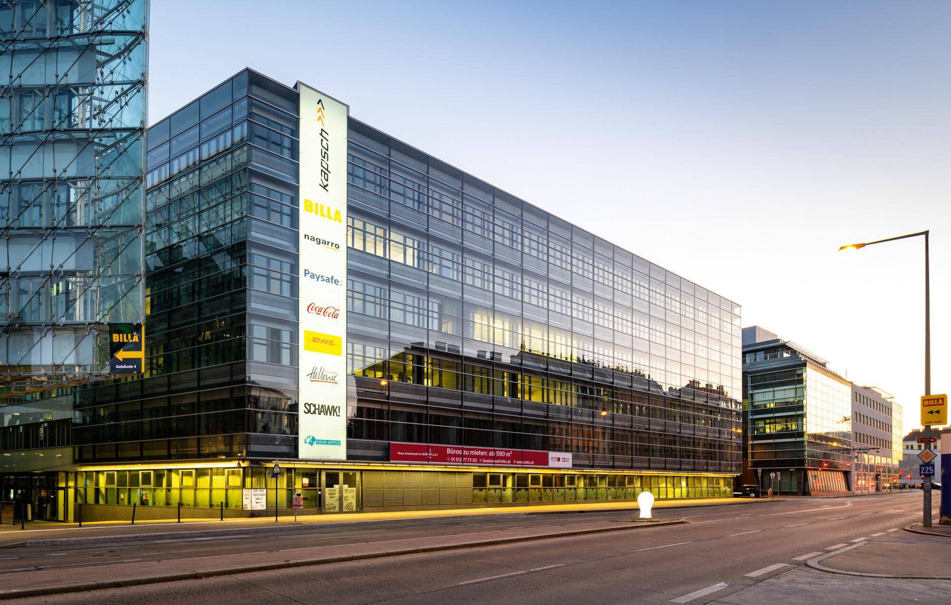 Modernes Bürogebäude mit Glasfassade und Werbeschildern an einer belebten Stadtstraße bei Tageslicht.