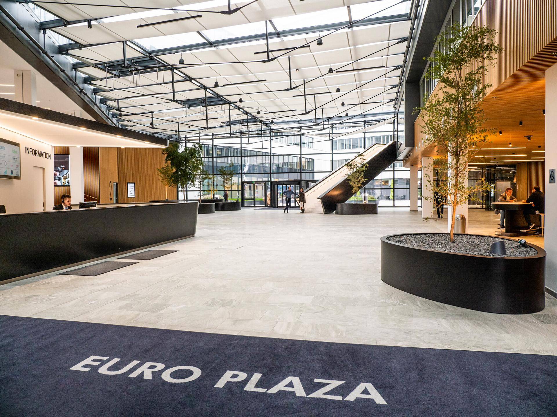 Geräumige, moderne Lobby mit Rezeption, Rolltreppe und großem Teppich mit Firmenlogo im Eingangsbereich.