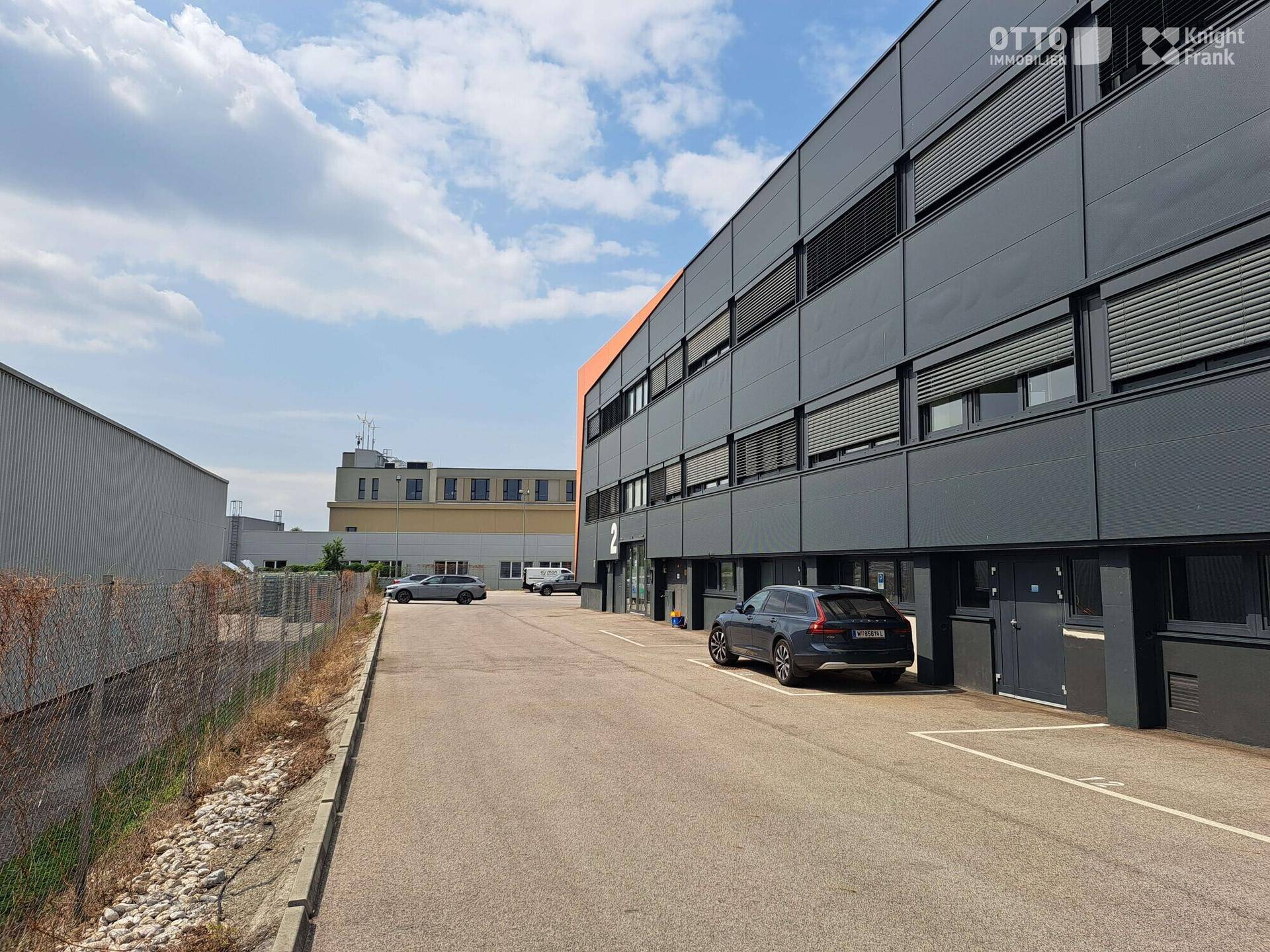Außenansicht eines modernen Büro- und Lagergebäudes mit Parkplätzen und Zufahrt.