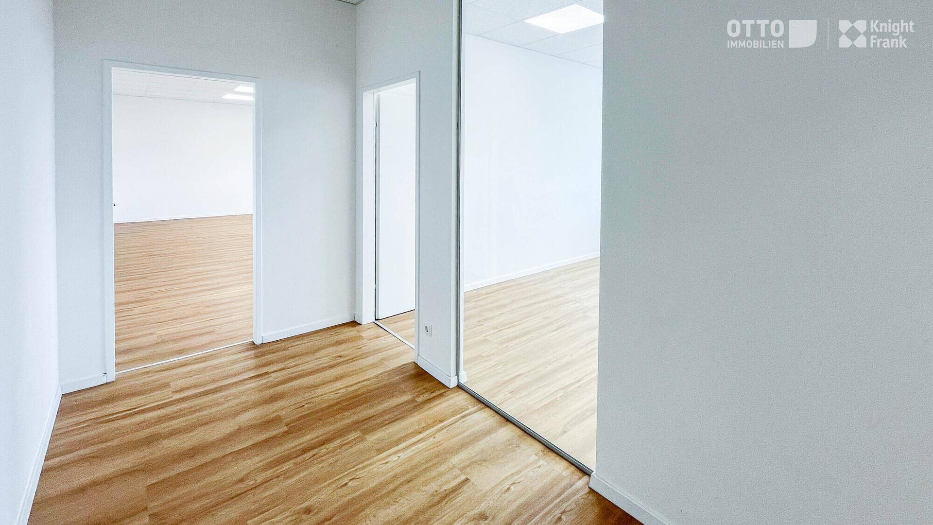 Heller Büroflur mit Holzoptikboden, weißen Wänden und modernen Glasabtrennungen zu angrenzenden Räumen.