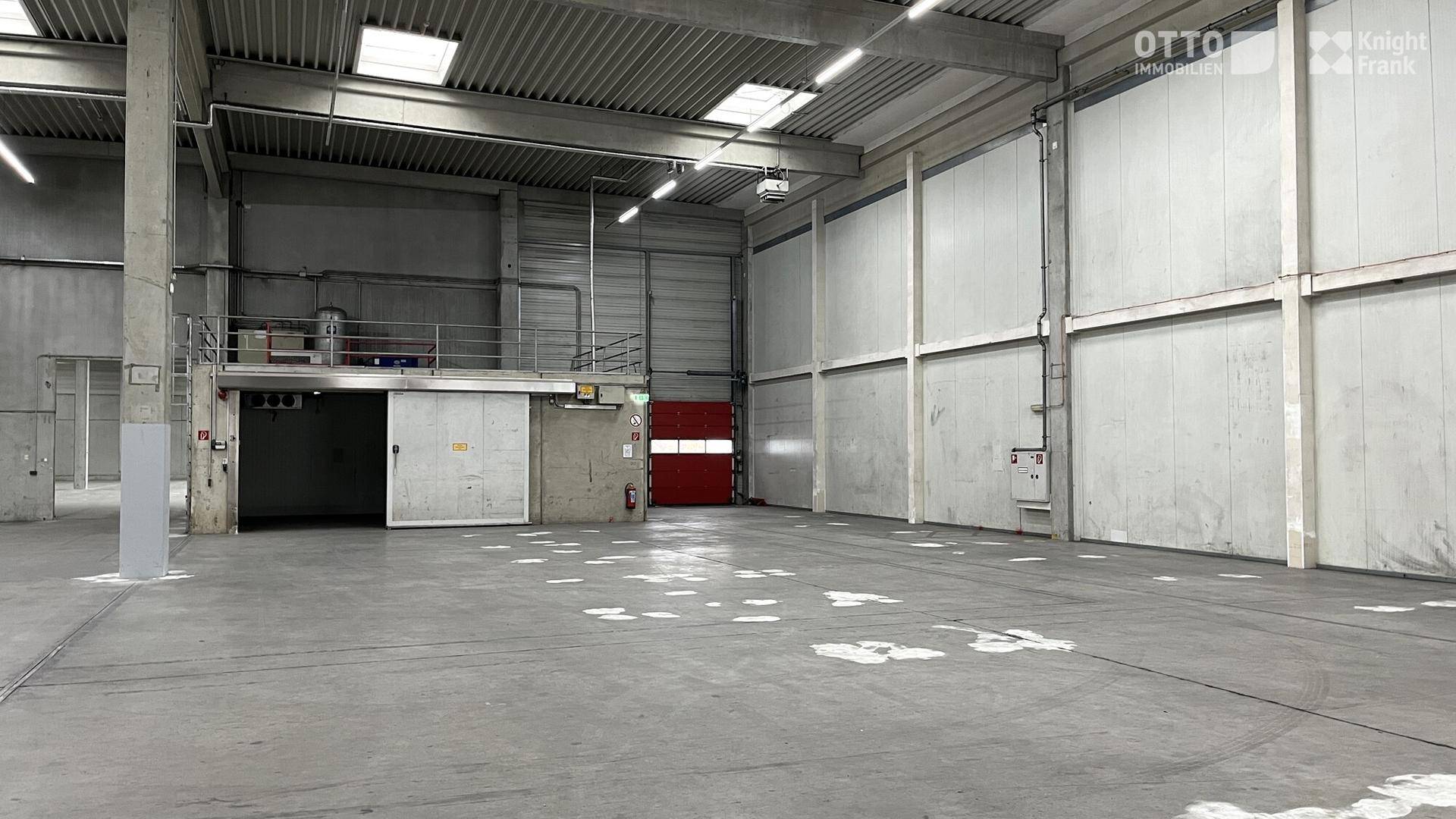 Leere Lagerhalle mit hohen Wänden, Betonboden und einem erhöhten Bereich im Hintergrund.
