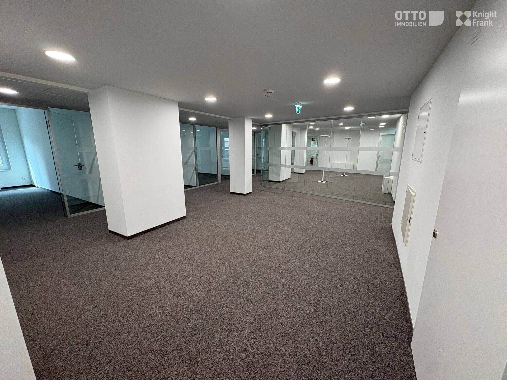 Offener Bürobereich mit Teppichboden, weißen Säulen und verglasten Büros im Hintergrund.