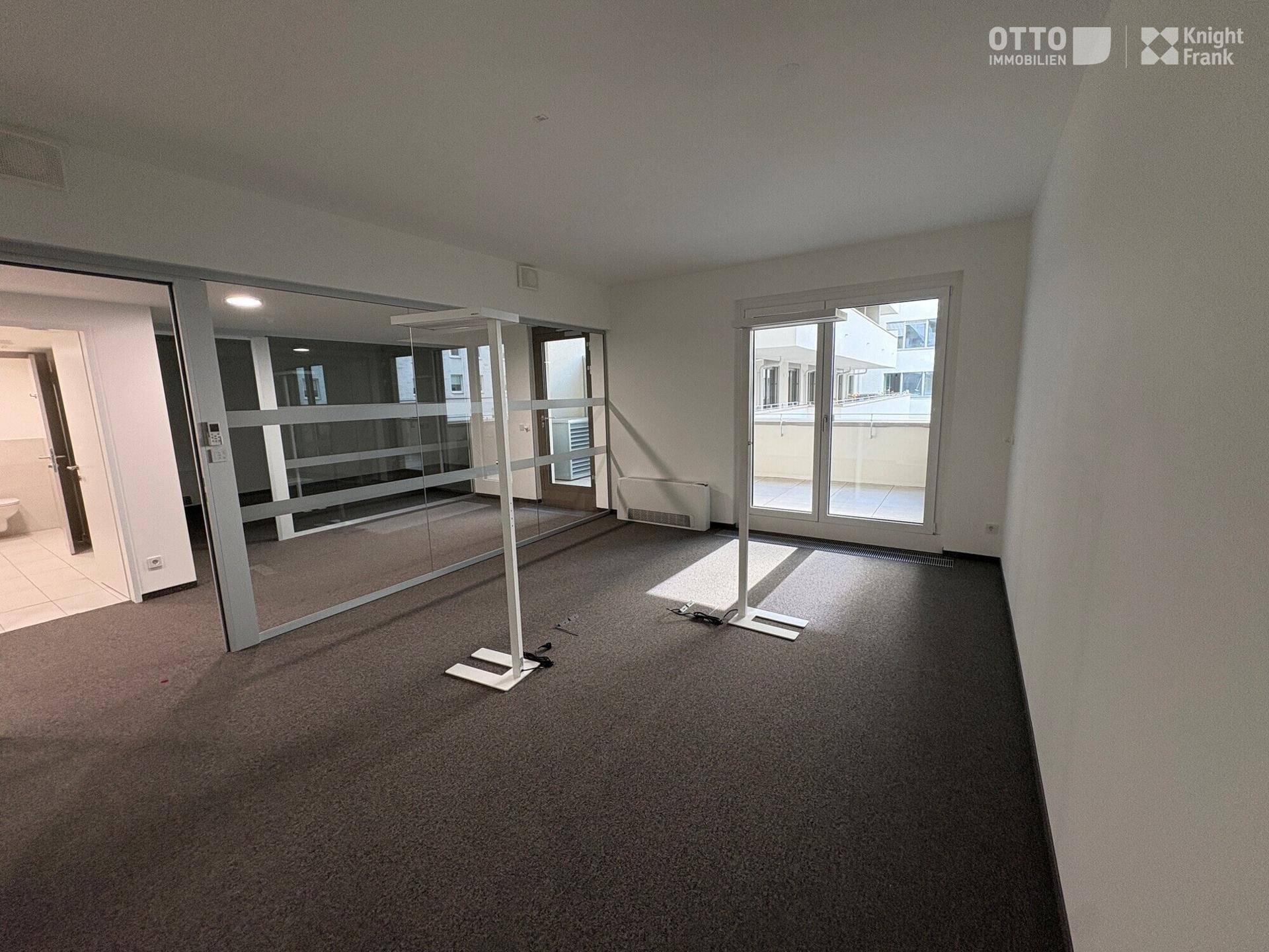 Heller Büroraum mit Teppichboden und großen Fenstern, die Zugang zu einem Balkon bieten.