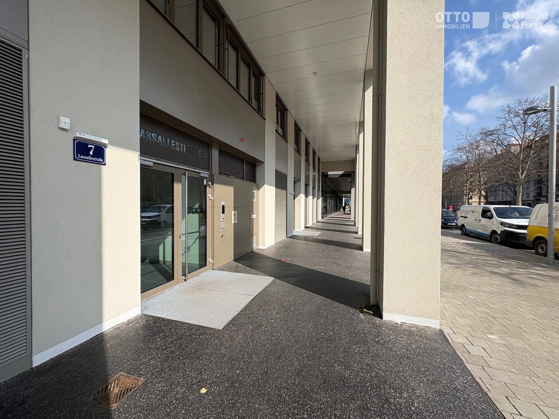 Eingangsbereich eines modernen Bürogebäudes mit breitem Gehweg und Säulen, unter blauem Himmel.